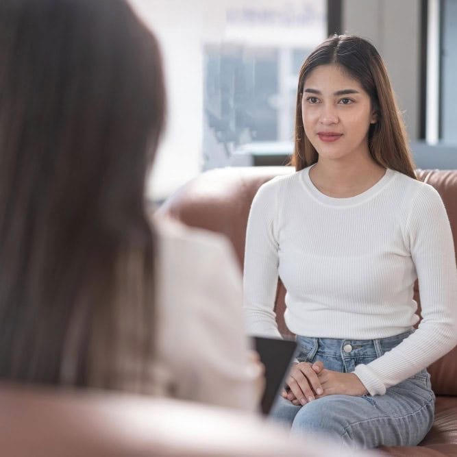 A young woman in a white long-sleeve shirt and jeans sits on a couch, attentively listening to another person whose back is to the camera in a bright, modern office setting. - KindestMind