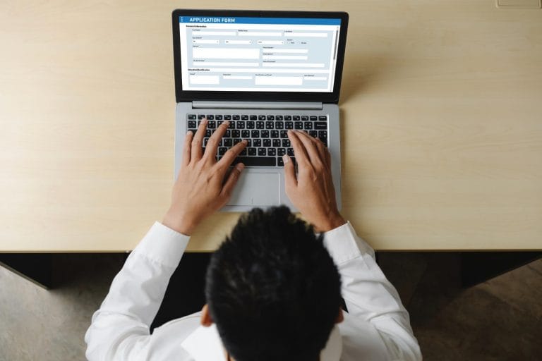 Our Process For Psychological And Neuropsychological Evaluations 1 A person in a white shirt is typing on a laptop at a wooden desk, filling out an online immigration evaluation form. The photo is taken from above, showing their hands and part of their head as they begin the evaluation process. - KindestMind
