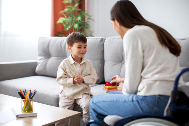 A young boy sits on a couch smiling at a woman in a wheelchair, who holds colorful stacking blocks—part of his autism evaluation. They are in a cozy living room with a table holding colored pencils in the foreground. - KindestMind