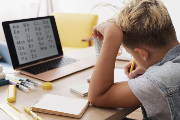 A young boy with blond hair writes in a notebook at a desk, while a laptop displaying the alphabet is open in front of him. Despite his diagnosed learning difference, he is thriving among scattered school supplies. - KindestMind