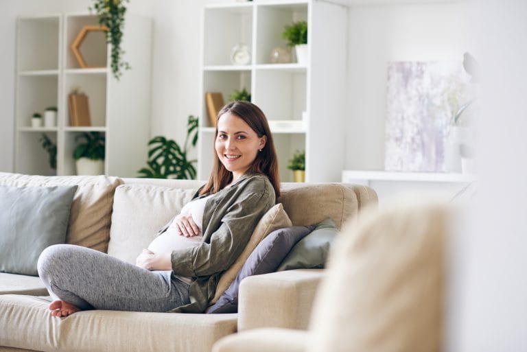 A pregnant woman sits comfortably on a beige sofa, smiling at the camera. Reflecting the psychological impact of surrogacy, she appears relaxed in a bright living room, decorated with plants and shelves, wearing a grey top and leggings with one hand on her belly. - KindestMind
