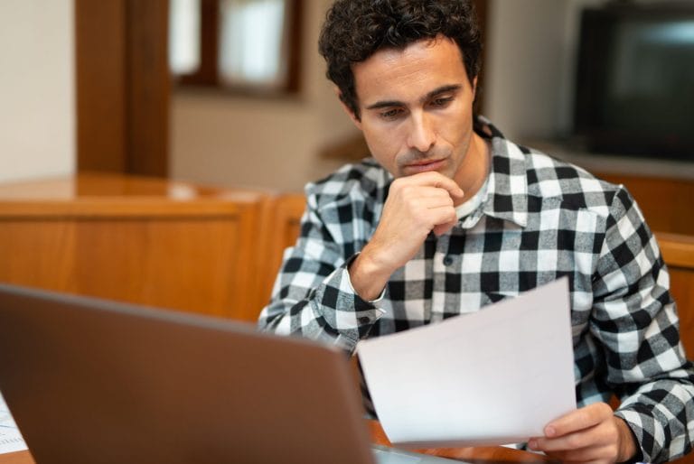 Our Process For Psychological And Neuropsychological Evaluations 5 A man wearing a black and white checkered shirt sits at a desk, looking thoughtfully at a sheet of paper while using a laptop, possibly reviewing documents for the immigration evaluation process. - KindestMind