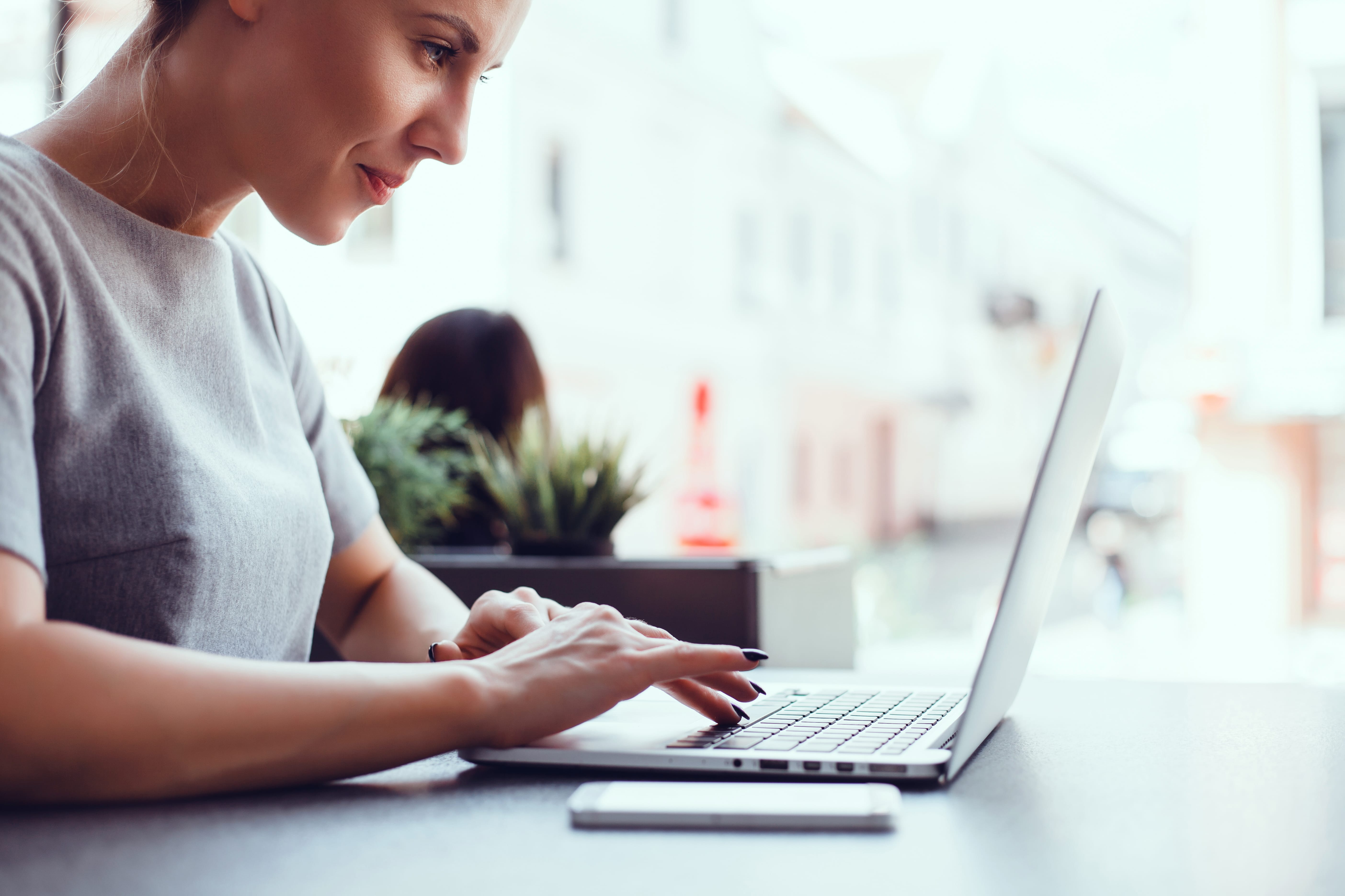 Our Process For Psychological And Neuropsychological Evaluations 2 A woman sits at a table in a bright indoor space, working on her laptop. She appears focused, perhaps reviewing psychological evaluations, with a smartphone beside her and potted plants in the background. - KindestMind
