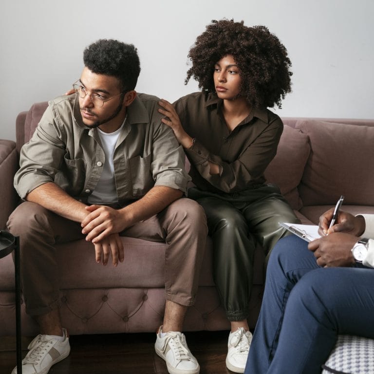 A woman sits on a couch comforting a concerned man by placing a hand on his shoulder. Across from them, someone with a notepad suggests theyre undergoing psychological evaluations during a counseling or therapy session. - KindestMind