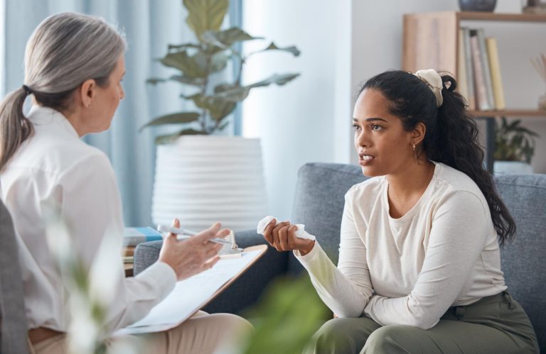 A young woman sits on a couch holding a tissue, talking to an older woman with a clipboard—suggesting post-surrogacy support in a comfortable, well-lit counseling office. - KindestMind