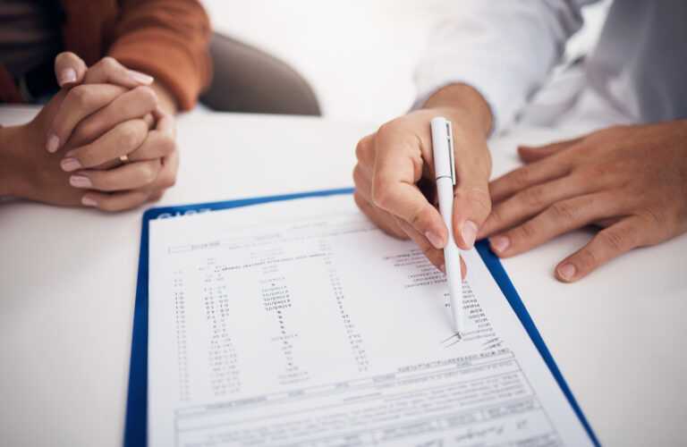 Two people sit at a desk, one holding a pen and pointing at a document on a clipboard while the other has their hands clasped, suggesting a discussion about the difference between psychological testing and neuropsychological evaluation. - KindestMind