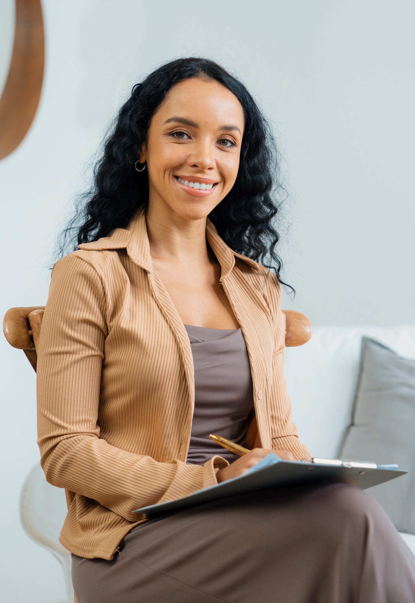 A woman with long curly dark hair, wearing a tan jacket and brown dress, sits on a chair indoors, smiling at the camera while holding a clipboard and pen—ready to assist with Immigration Psychological Evaluations at KindestMind. - KindestMind