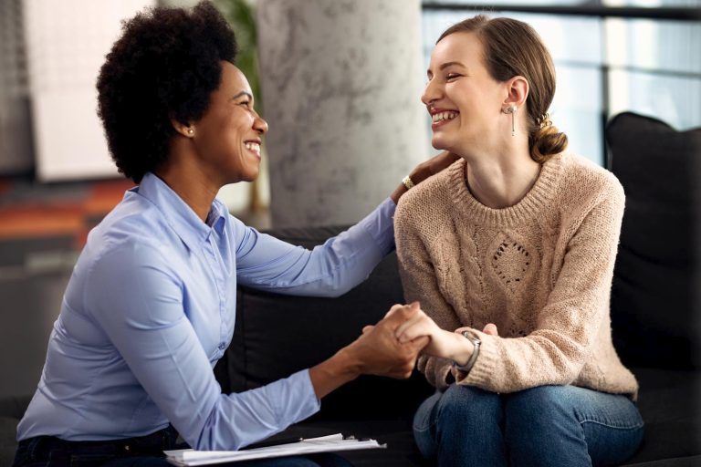 Two women sit on a couch, smiling warmly and holding hands. One holds a clipboard, the other wears a tan sweater and jeans. They appear to be having a supportive conversation about their surrogacy journey and seeking support together. - KindestMind