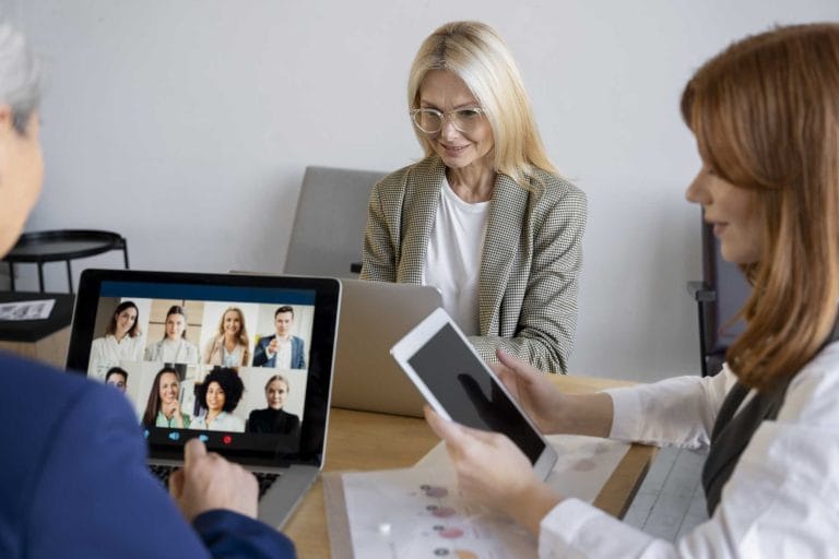 Three women work at a table with laptops and a tablet. One woman joins a video call for staff training, while the others focus on their own devices and documents, supporting tasks at a surrogacy agency. - KindestMind