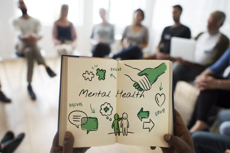 A person holds an open notebook showing doodles and the words Mental Health. In the background, a group sits in a circle, suggesting a support group or therapy session focused on well-being and open discussion. - KindestMind