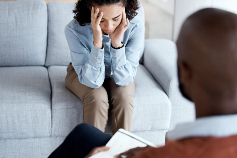 A woman sits on a couch, distressed, her head in her hands. In the foreground, a person with a notepad listens attentively, suggesting a counseling session focused on compassionate care and trauma-informed evaluation. - KindestMind