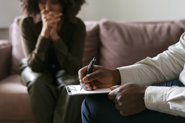 A person sits with hands clasped, looking concerned, while another takes notes on a clipboard, suggesting psychological evaluations and highlighting the benefits for domestic abuse survivors in a therapy session. - KindestMind
