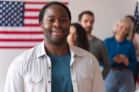 A smiling man stands at the front of a line of diverse people, with a U.S. flag hanging on the wall behind them. The group appears to be waiting, possibly for a legal immigration event or to become a U.S. citizen. - KindestMind