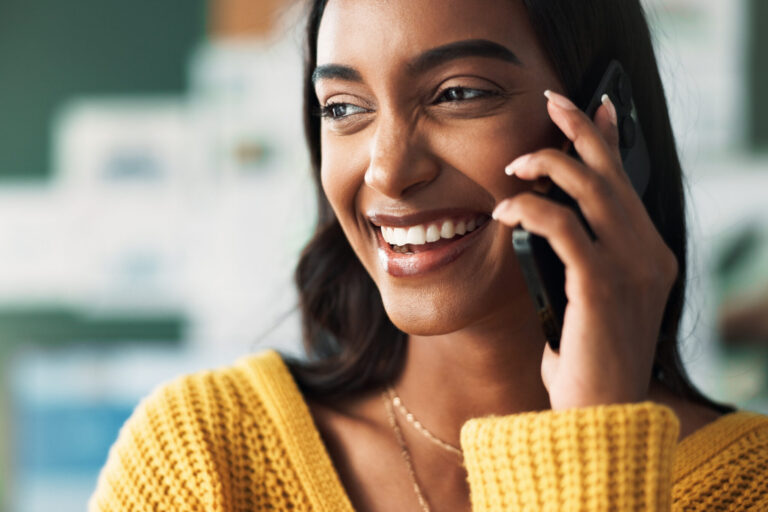 A woman wearing a yellow sweater smiles while talking on a smartphone, with a blurred background suggesting an indoor setting. - KindestMind
