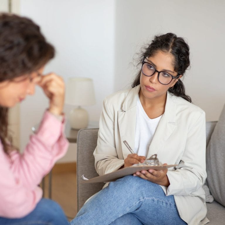 A therapist conducts Psychological Evaluations, listening attentively and taking notes as a distressed woman seeking asylum sits across from her, holding her forehead in a calm, indoor setting with neutral decor. - KindestMind