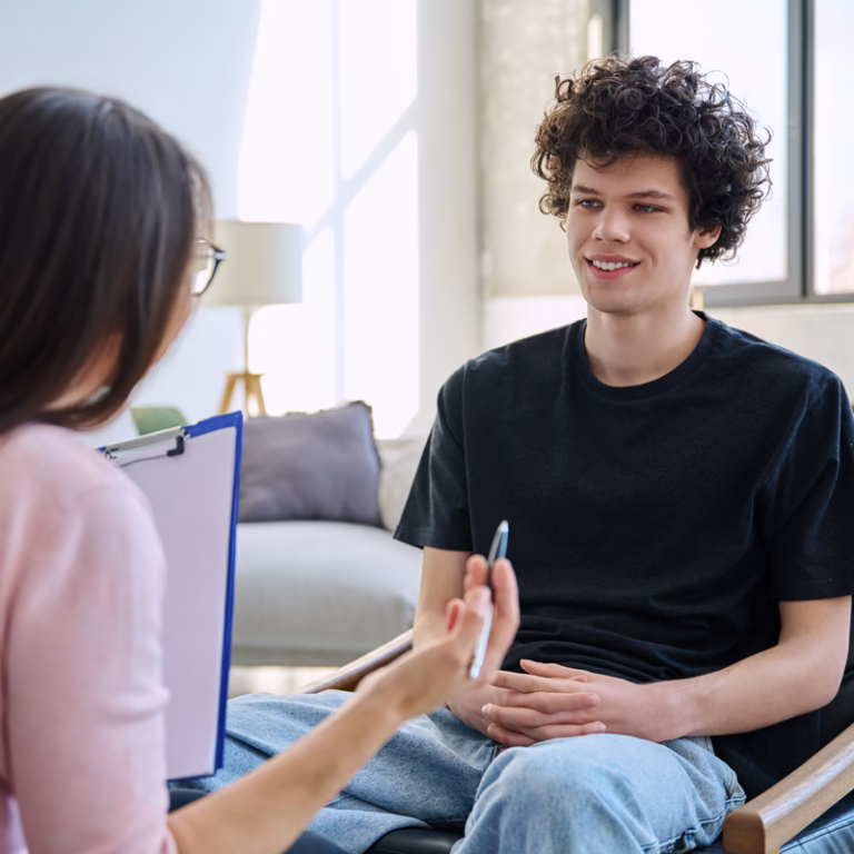 A young man with curly hair smiles while sitting across from a woman holding a clipboard and pen during assessments about substance use in a bright, modern room. - KindestMind