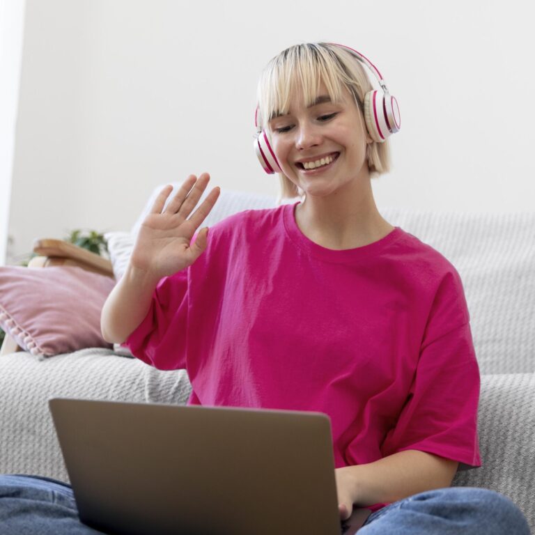 A smiling person wearing headphones and a bright pink shirt waves at their laptop while sitting on a couch, appearing to be on a video call in a cozy living room for standard Autism Spectrum Disorder assessments. - KindestMind