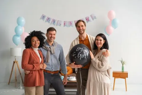 Four smiling adults stand together at a gender reveal party, holding a black balloon that reads Boy or Girl? Pink and blue decorations fill the background, celebrating their journey with independent surrogacy. - KindestMind