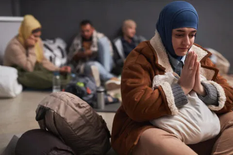 A woman wearing a blue hijab and brown coat sits on the floor, clasping her hands in prayer. Behind her, three others with luggage look worried and tired, capturing the emotional toll of an asylum case or pending psychological evaluation. - KindestMind