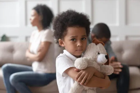 A young child hugs a teddy bear and looks sad, standing in the foreground. In the background, two adults sit on a sofa facing away from each other, appearing upset—a scene often linked to childhood trauma and its impact on brain development. - KindestMind