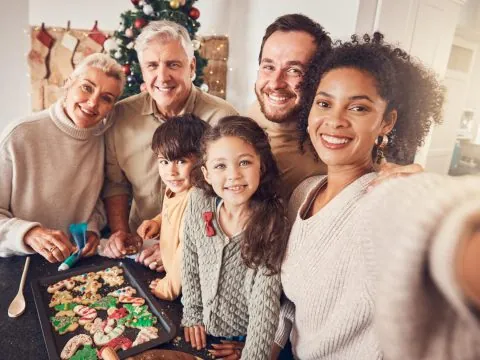 A multigenerational family smiles for a selfie in a festive kitchen, gathered around a tray of decorated holiday cookies, celebrating the holidays together with a Christmas tree and stockings in the background. - KindestMind