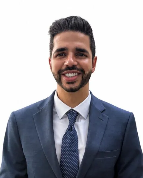 A member of the Clinical Team, a man in a navy suit, white shirt, and striped tie smiles at the camera against a plain white background. - KindestMind