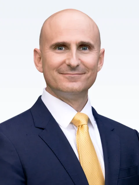 A man in a navy blue suit with a white shirt and yellow tie, smiling and standing against a plain light background, ready to support the Clinical Team. - KindestMind