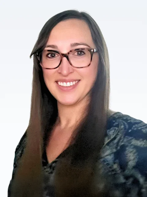 A woman with long brown hair wearing glasses and a patterned top smiles at the camera against a plain light background, reflecting the warmth and approachability of our Clinical Team. - KindestMind