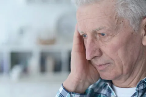 An elderly man with light skin and gray hair, wearing a plaid shirt, holds his hand to the side of his face and looks thoughtful or concerned in a softly lit indoor setting, perhaps reflecting on his legal capacity or personal autonomy. - KindestMind