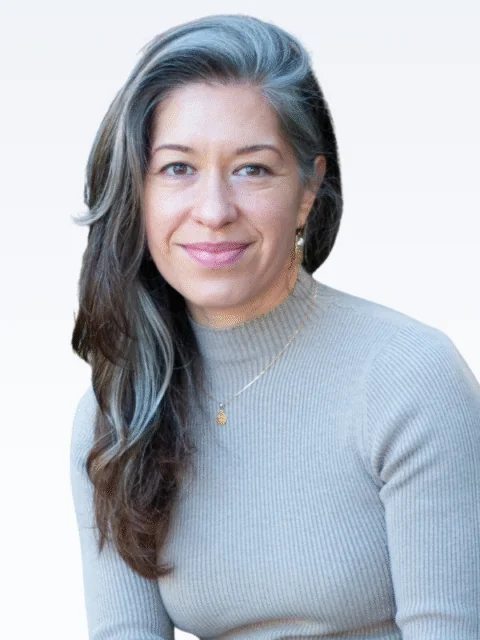 A woman with long, wavy brown and gray hair, wearing a beige ribbed turtleneck sweater and gold jewelry, smiles at the camera against a light gray background, capturing the approachable spirit of our Clinical Team. - KindestMind
