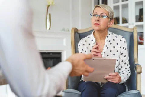 A woman with short blond hair and glasses sits in a chair, holding a clipboard and pen, attentively listening during a forensic psychological evaluation with a person whose hands are visible in the foreground. - KindestMind