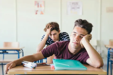 Two teenage students sit at desks in a classroom, looking bored and tired—common ADHD symptoms. One boy rests his head on his hand, while the other has his head down behind him. School supplies are scattered on their desks. - KindestMind