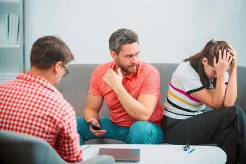 Three people sit on a couch during a counseling session addressing surrogacy conflicts; a man in red looks concerned, a woman holds her head in distress, and another man in glasses takes notes. Glasses and a folder are on the table in front of them. - KindestMind