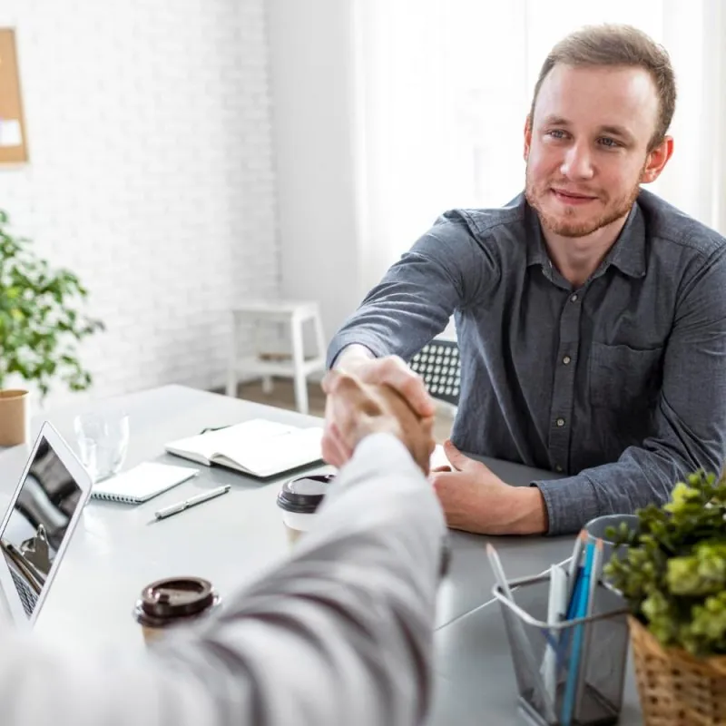 A man in a blue shirt smiles and shakes hands with another person across a desk in a modern office, suggesting a positive meeting about Disability Accommodation. Office supplies, laptops, and plants are visible on the desk. - KindestMind