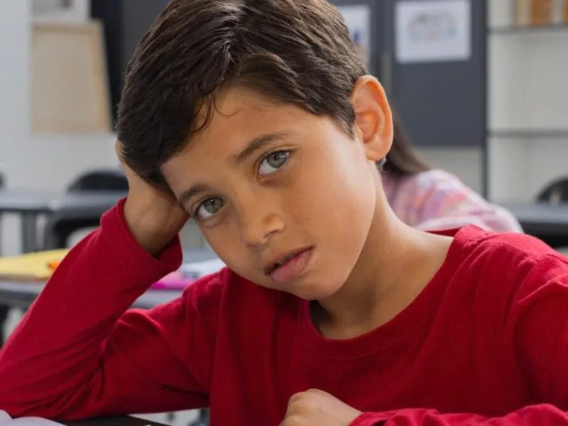 A young boy with light skin and brown hair, wearing a red shirt, sits at a desk in a classroom, leaning his head on one hand and looking thoughtfully at the camera—perhaps during diagnostic assessments for ADHD—with notebooks and a tablet in front of him. - KindestMind