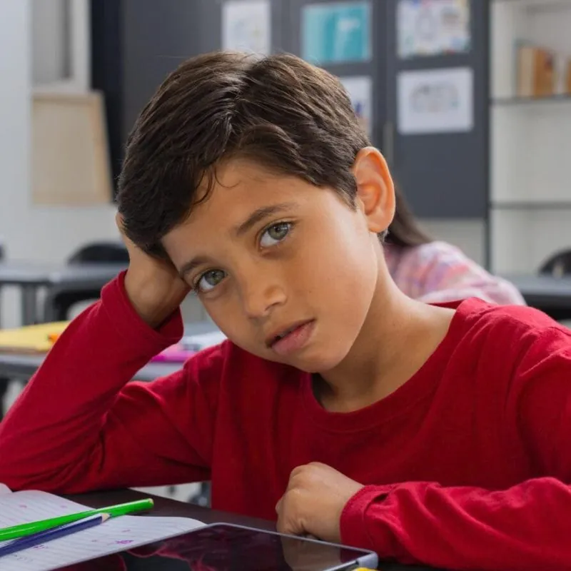 A young boy with light skin and brown hair, wearing a red shirt, sits at a desk in a classroom, leaning his head on one hand and looking thoughtfully at the camera—perhaps during diagnostic assessments for ADHD—with notebooks and a tablet in front of him. - KindestMind