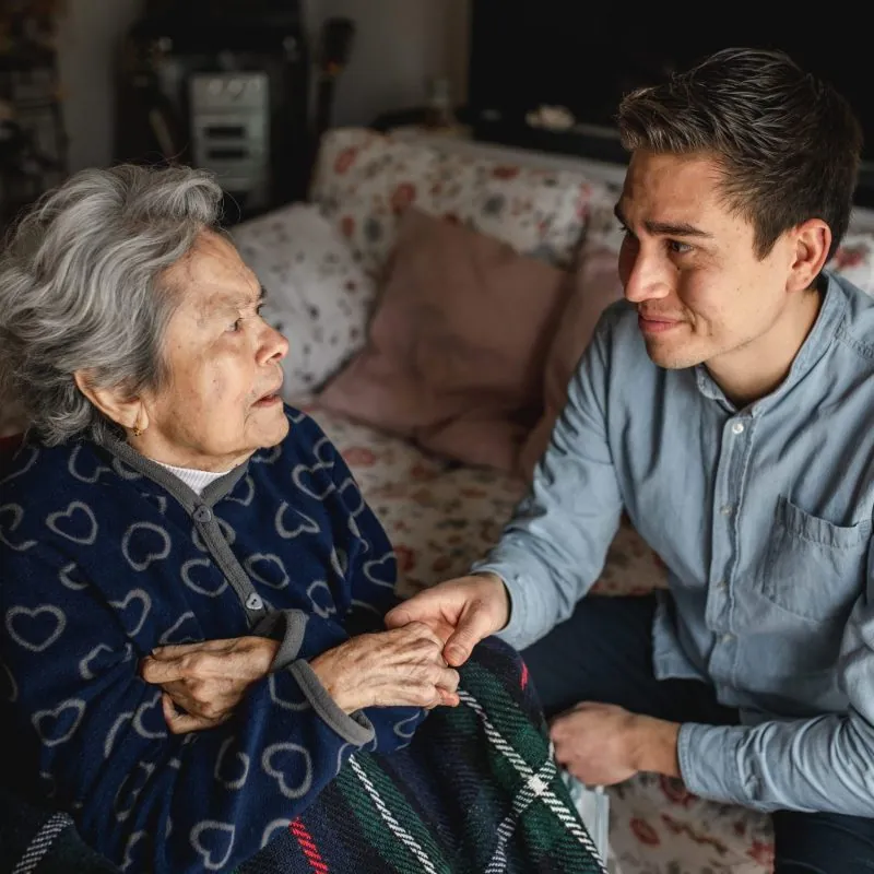 A young man holding hands and comforting an elderly woman sitting on a couch with a blanket, sharing a caring and emotional moment together in a cozy living room—capturing the support often needed in extreme hardship waiver or I-601 cases. - KindestMind