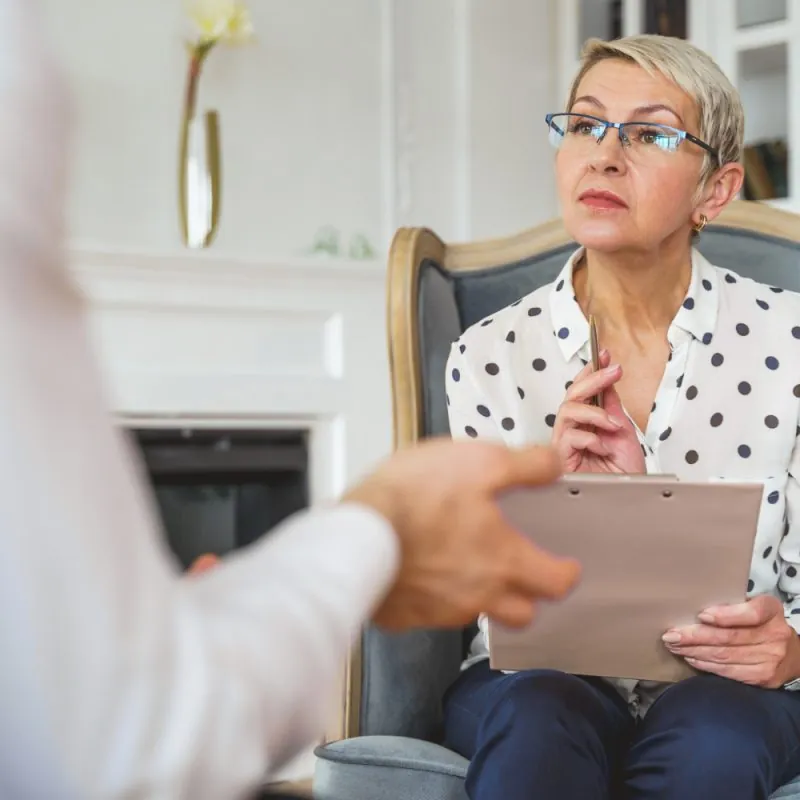 A woman with short blond hair and glasses sits in a chair, holding a clipboard and pen, attentively listening during a forensic psychological evaluation with a person whose hands are visible in the foreground. - KindestMind