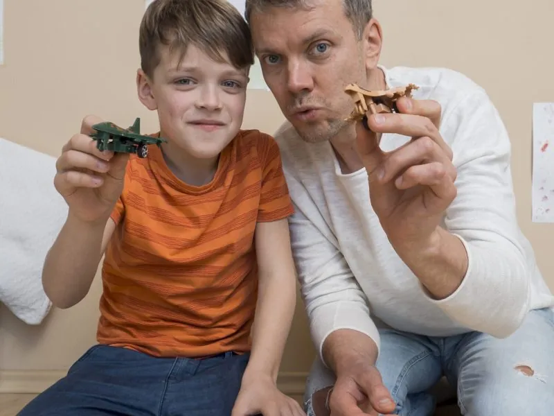 A man and a boy sit on the floor indoors, smiling and holding toy airplanes toward the camera. Comprehensive assessments for Autism Spectrum Disorder can help support joyful moments like these. Drawings are visible on the wall behind them. - KindestMind