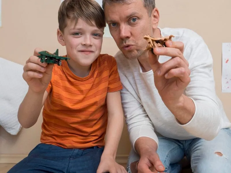 A man and a boy sit on the floor indoors, smiling and holding toy airplanes toward the camera. Comprehensive assessments for Autism Spectrum Disorder can help support joyful moments like these. Drawings are visible on the wall behind them. - KindestMind