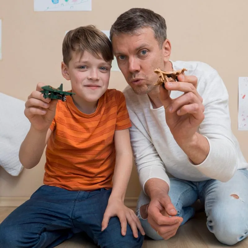 A man and a boy sit on the floor indoors, smiling and holding toy airplanes toward the camera. Comprehensive assessments for Autism Spectrum Disorder can help support joyful moments like these. Drawings are visible on the wall behind them. - KindestMind