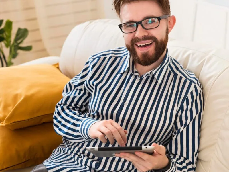 A smiling man with glasses and a striped shirt sits on a white couch, holding and using a tablet—perhaps completing assessments related to Autism Spectrum Disorder. Yellow pillows are behind him, with a plant in the background. - KindestMind