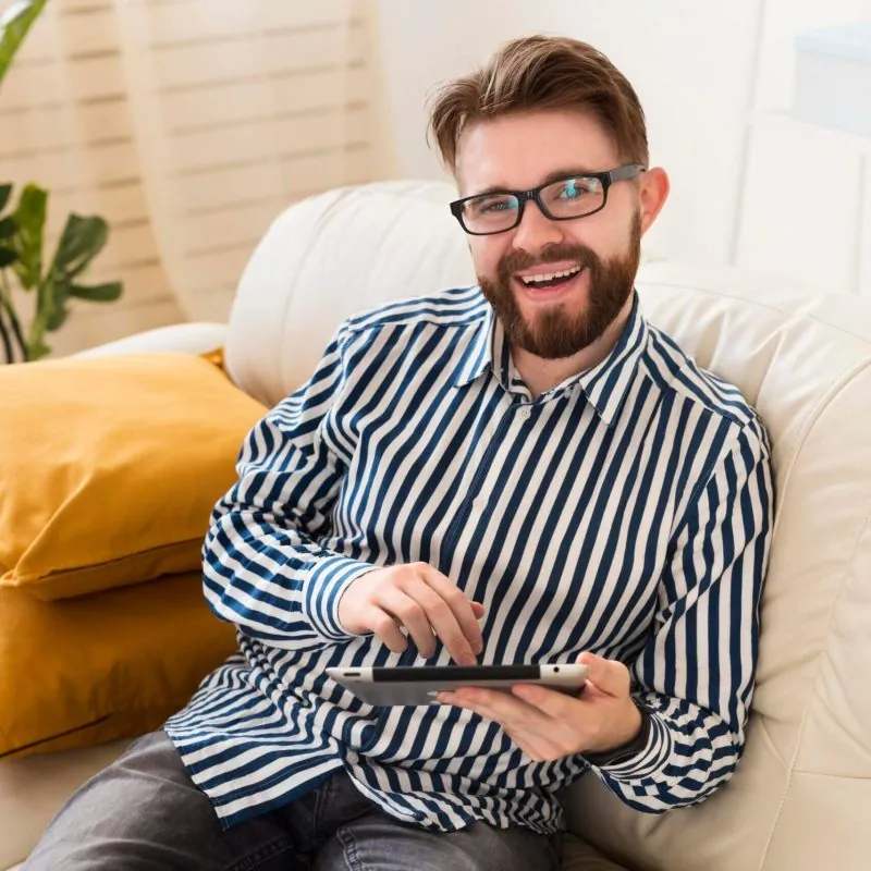 A smiling man with glasses and a striped shirt sits on a white couch, holding and using a tablet—perhaps completing assessments related to Autism Spectrum Disorder. Yellow pillows are behind him, with a plant in the background. - KindestMind