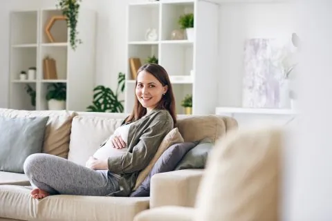 A pregnant woman sits comfortably on a beige sofa, smiling at the camera. Reflecting the psychological impact of surrogacy, she appears relaxed in a bright living room, decorated with plants and shelves, wearing a grey top and leggings with one hand on her belly. - KindestMind