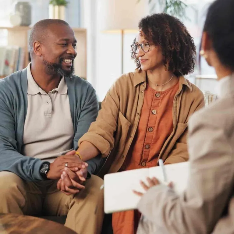 A couple sits close together holding hands and smiling warmly at each other during a counseling session with a therapist who is taking notes. - KindestMind
